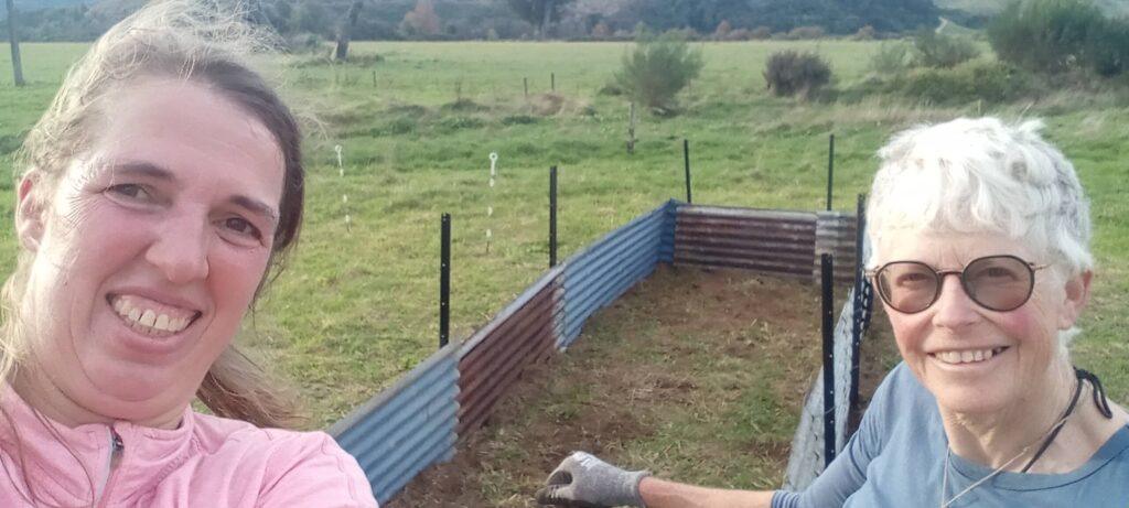 Two women smiling proudly in front of a newly built corrugated iron raised garden bed at Bainham Hall