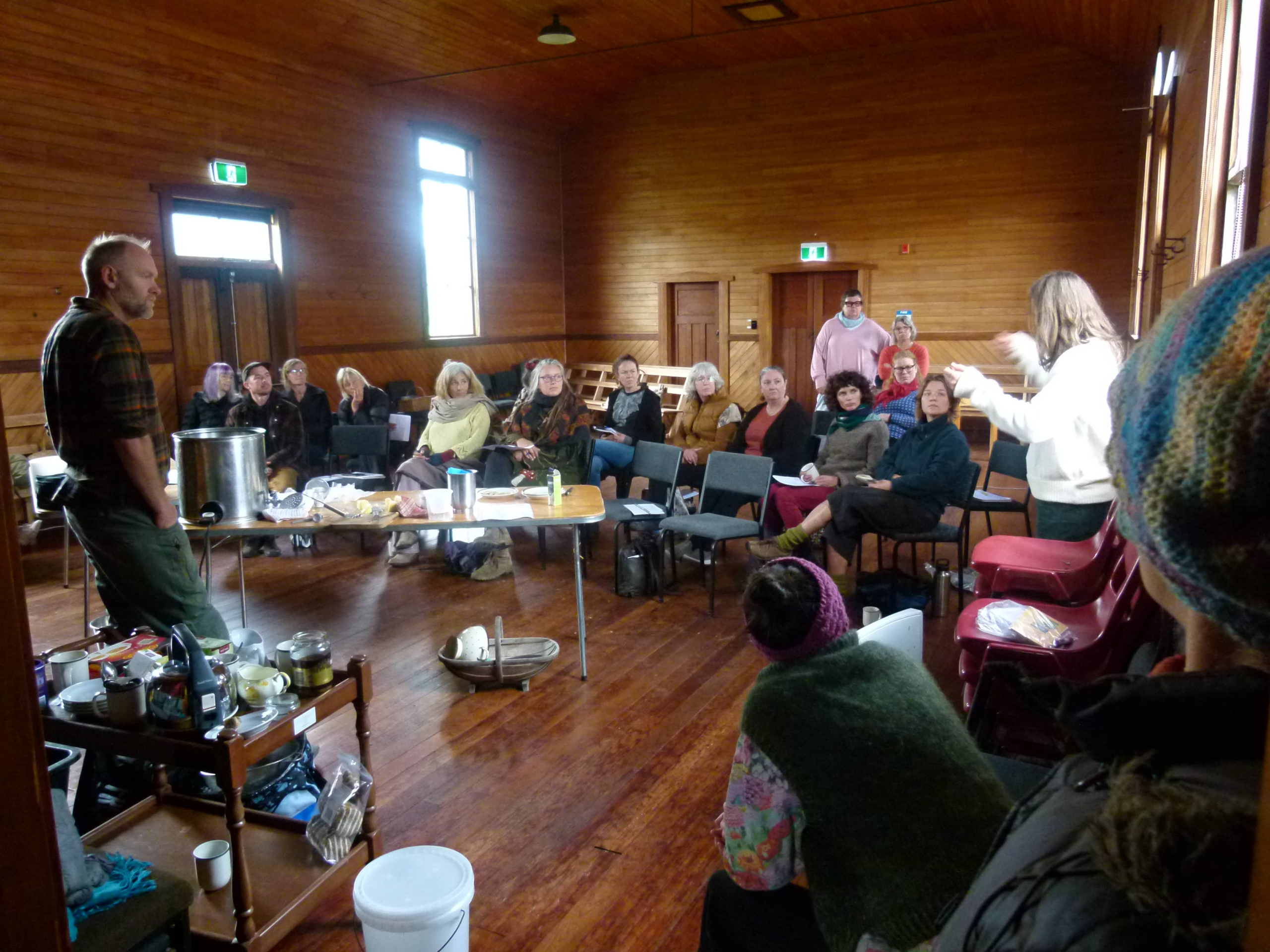 Two tutors at the front of Bainham Hall demonstrating cheese making to a large seated audience at the Aorere Valley Practical Living Series.