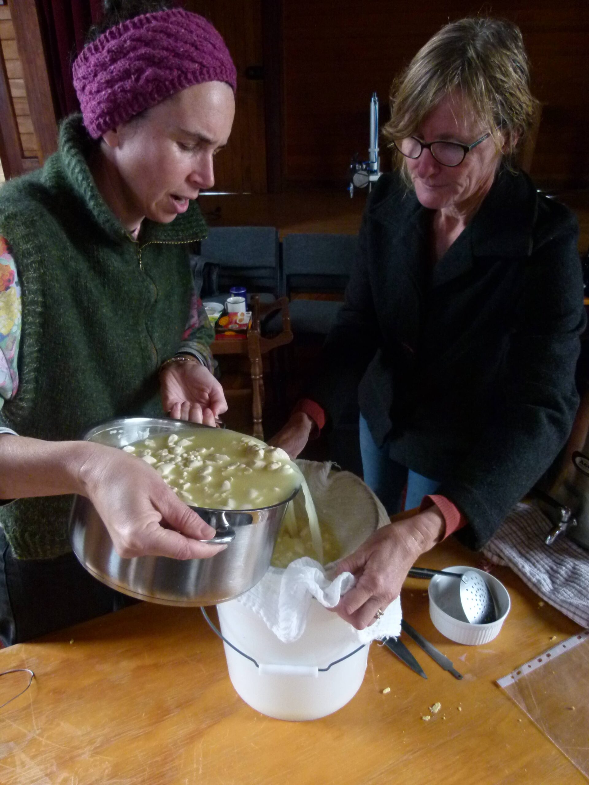 Two women carefully pour mozzarella curds from a metal bowl through cheesecloth into a colander at a wooden table at Bainham Hall