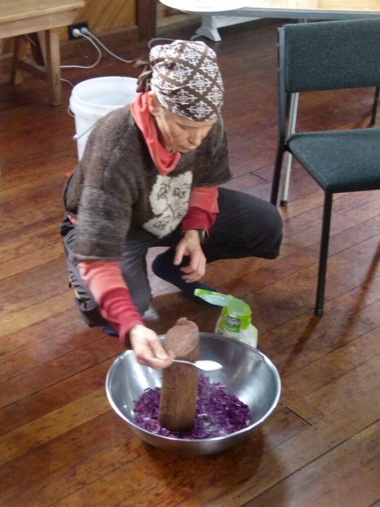 A woman kneeling on a wooden hall floor, pounding shredded purple cabbage with salt in a large metal bowl using a wooden pounder.