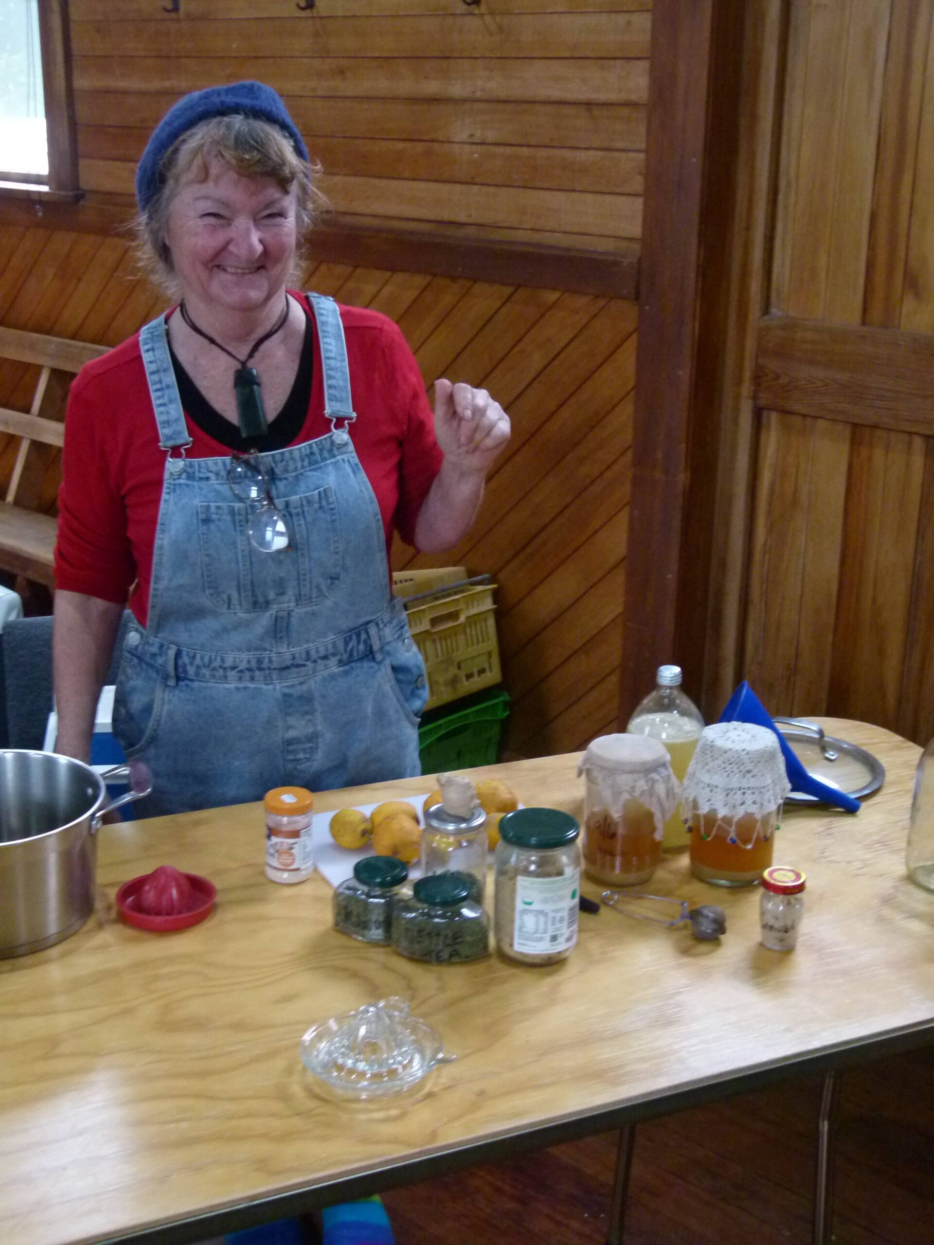 A smiling woman in denim overalls standing behind a table set up with kombucha jars, a ginger beer bottle, lemons, tea, sugar, and fermenting supplies.