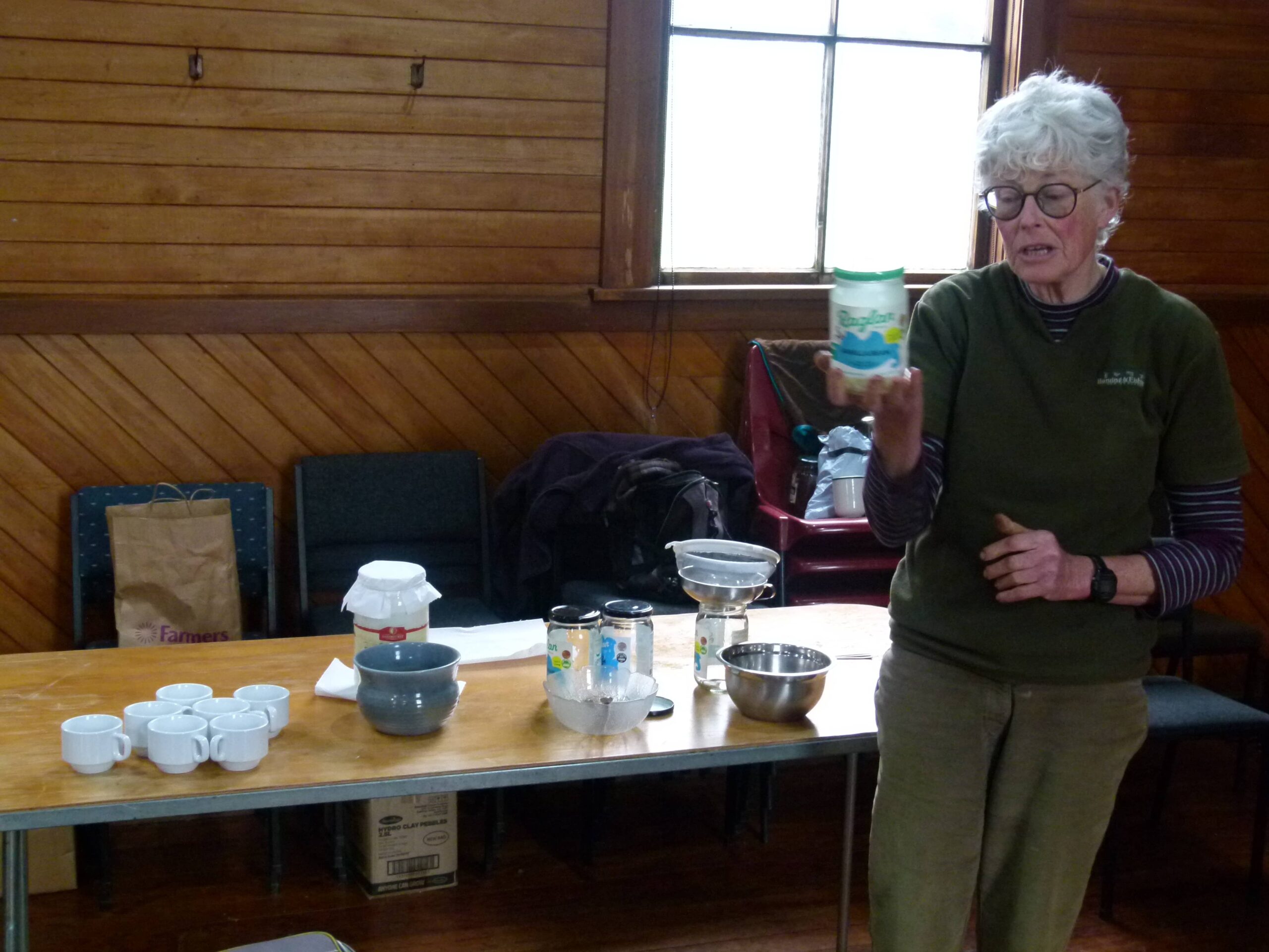 A woman holding up a container while explaining dairy kefir, with cups, jars, and a strainer set out on a table in front of her.